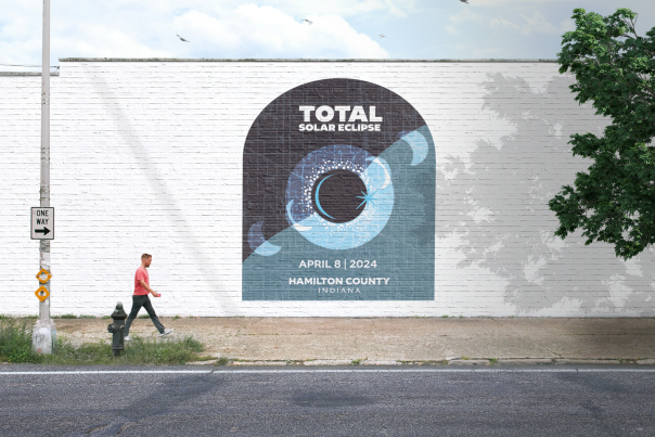 Large mural on a white brick wall depicting a stylized total solar eclipse with radiating blue arcs and text reading ‘Total Solar Eclipse — April 8, 2024 — Hamilton County, Indiana,’ with a person walking past on the sidewalk.