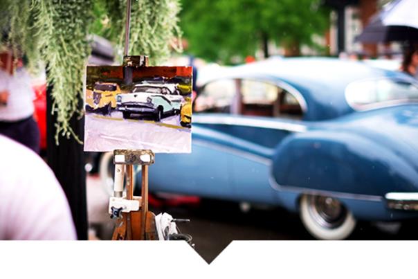 Close-up of a small framed photograph clipped to a wooden display, with a light blue vintage car parked in the background. People and market-style booths appear softly blurred behind the car, suggesting an outdoor event.