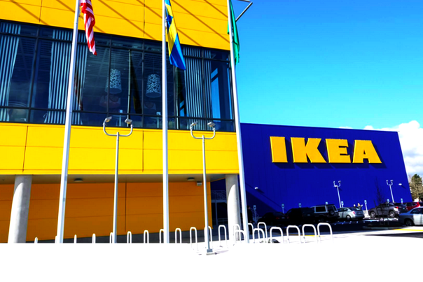Exterior of an IKEA store with a bright blue facade and large yellow “IKEA” lettering. Yellow and blue building sections rise behind several flagpoles, with a parking area and shopping carts visible in front under a clear blue sky.