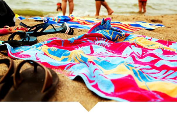 Colorful beach towels spread out on sand near the water, with sandals and personal items resting on top. People’s legs are visible in the background near the shoreline.