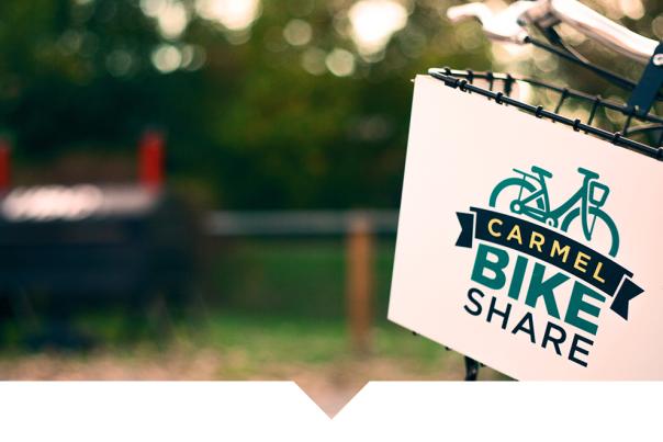 A white rectangular sign hanging from a metal bracket on a bicycle, displaying a teal bicycle graphic above bold text that reads “CARMEL BIKE SHARE.” The background is softly blurred, showing greenery and an outdoor setting.