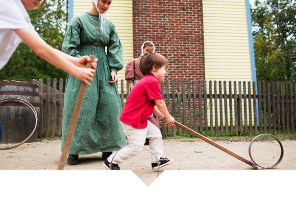 A child pulling a long-handled wooden tool across a dirt path while an adult in historical-style clothing stands nearby.