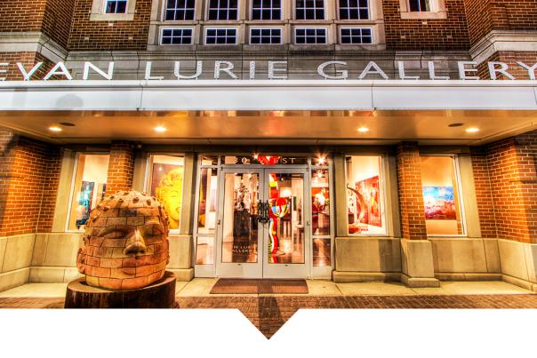 The entrance to a brick gallery building with a marquee that reads “EVAN LURIE GALLERY.” Glass double doors reveal artwork inside, and a sculpted stone head sits on a pedestal in front of the entrance.