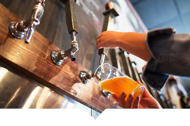 Beer being poured from metal taps into a clear glass at a wooden bar.