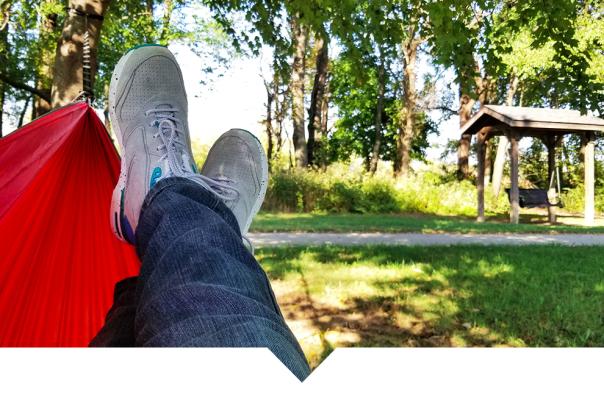 A first-person view of legs in blue jeans and gray sneakers resting in a red hammock in a grassy park.