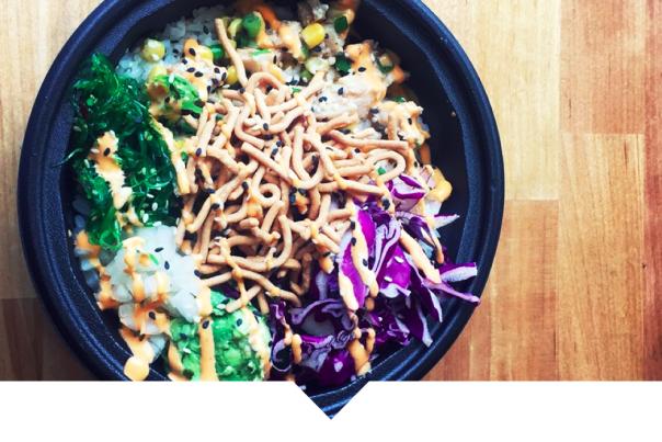 Poke bowl in a black bowl filled with vegetables, noodles, and sauce, set on a wooden tabletop.