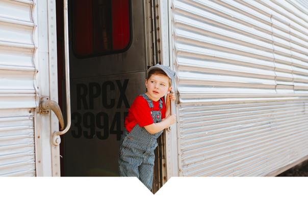 A young child wearing denim overalls and a grey cap stands in the open doorway of a silver vintage train car.