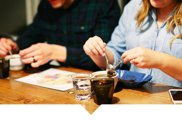 Two people seated at a wooden table, preparing coffee with small cups, tools, and water glasses.