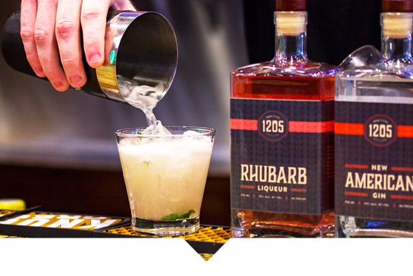 A close-up of a drink being poured into a glass beside bottles labeled “Rhubarb” and "American."