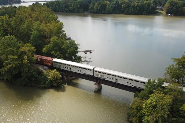 An overhead view of a passenger train crossing a bridge over a wide river, with tree-lined banks on both sides. The train stretches across the water, surrounded by dense green foliage.
