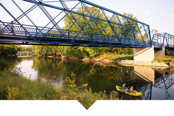 A blue steel truss bridge spans a calm river, reflecting in the water below. Trees and grassy banks line the river, with a small group in a kayak visible near the shore.