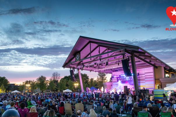 An outdoor amphitheater stage illuminated in purple light with a crowd gathered in front at dusk and a red logo that reads "#LOVEHC."