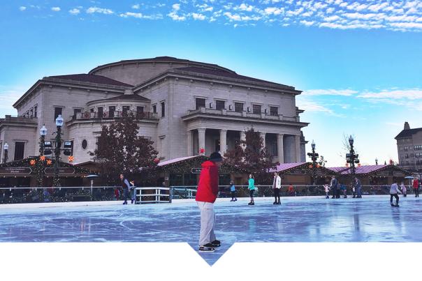 Outdoor ice skating rink set in front of a large neoclassical-style building with tall columns, under clear blue skies on a sunny day.