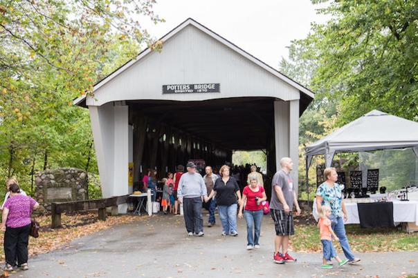 A group of people walking through a white wooden covered bridge with a sign above the entrance that reads "POTTER'S BRIDGE".