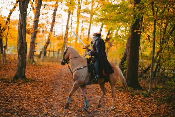 A headless person in a black cape, with a sword, riding a brown horse through a forest filled with orange and yellow autumn leaves.