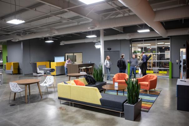 A modern remote work office space features an open floor plan with concrete floors and exposed ceilings. The room includes a mix of seating areas, such as sofas, lounge chairs, and shared tables, arranged for both collaboration and individual work. Bright accent furniture in yellow and orange adds color, while potted plants soften the industrial design. Several people stand and talk near a glass-walled meeting room in the background, creating a casual, flexible workplace atmosphere.