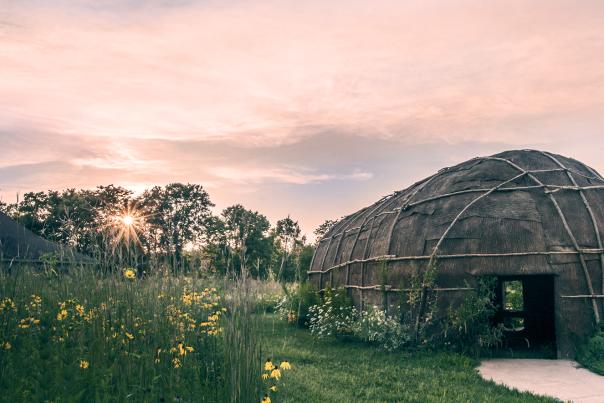 A hut building with a trail and surrounded by greenery, against a pink and purple sunset sky.