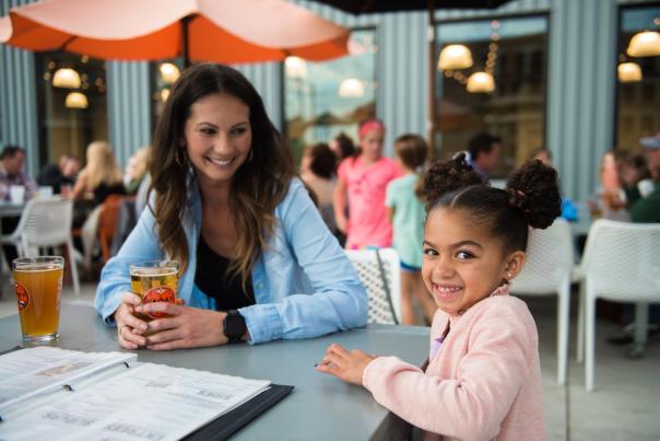 A woman and a young girl sit together at an outdoor restaurant table, smiling toward the camera, with drinks, menus, and other diners visible in the background.