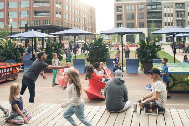 A lively urban street with people walking, children playing, outdoor dining, and brick buildings
