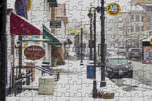 A jigsaw puzzle image depicting a snowy main street with storefronts, parked cars, streetlights, and hanging signs, with puzzle piece outlines visible across the scene.