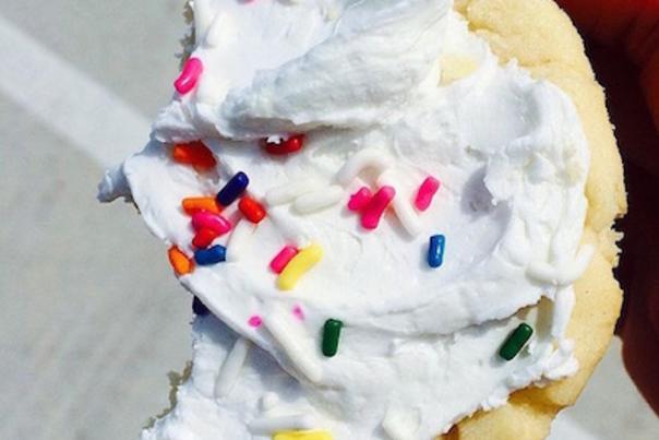 A white sugar cookie with white frosting and rainbow sprinkles, with a bite taken out of one side, is held in a hand above a paved parking lot surface.