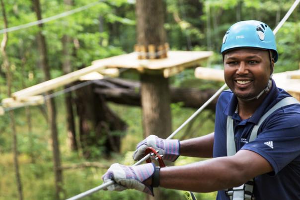 A person wearing a blue safety helmet and harness smiles while navigating an aerial ropes course among the trees.