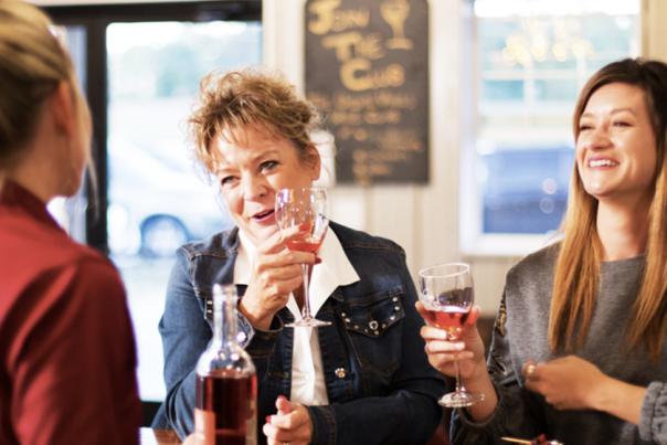 Three people seated at a table inside a café, holding glasses of wine and smiling or laughing. The indoor setting includes a small table and a casual dining atmosphere.