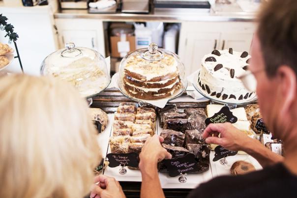 People look over a bakery counter displaying various treats, including a large frosted cake and several smaller pastries on white platters.