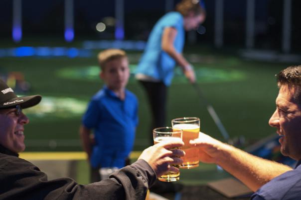 Two people clinking glasses of beer on a balcony overlooking a golf driving range where a child stands in the background.