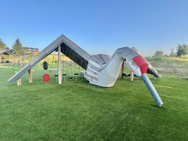 A large bird-shaped playground climbing structure sits on a grassy play area. The wooden structure features a long beak, red head, curved body, rope climbing elements, and suspended stepping platforms beneath peaked roof sections. Open green space, additional playground equipment, and trees are visible in the background under a clear blue sky.