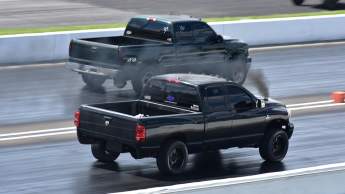 Two diesel pickup trucks race side by side on a drag strip, releasing black smoke as they accelerate.
