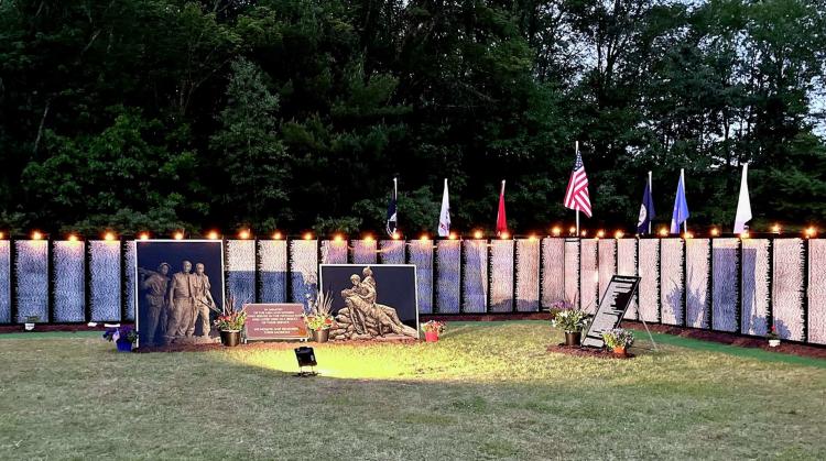 Vietnam Traveling Memorial Wall illuminated at dusk, with American and military flags flying above the memorial panels and tributes honoring those who served in the Vietnam War.