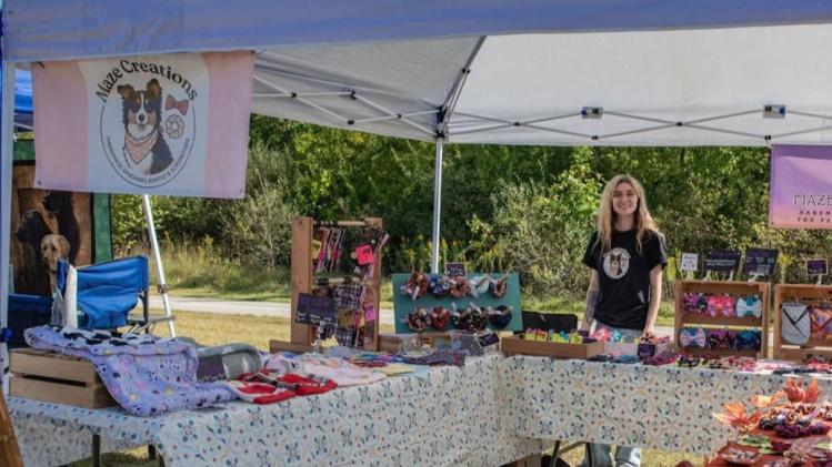 Woman stands behind vendor booth with a variety of dog supplies for sale.