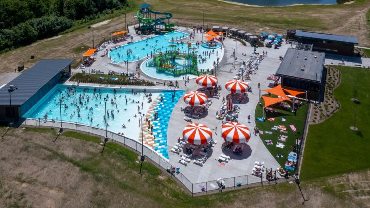Aerial view of a crowded outdoor aquatic center featuring pools, waterslides, splash play areas and striped shade umbrellas on a sunny summer day in Hendricks County, Indiana.
