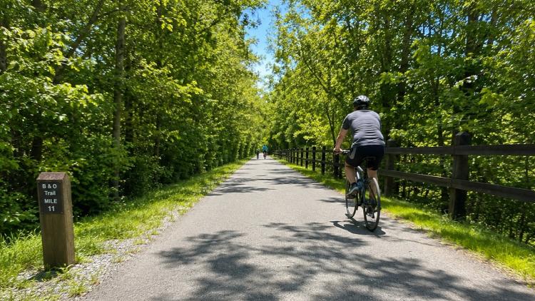 Cyclists travel along the paved B&O Trail through a wooded corridor on a sunny summer day in Brownsburg.