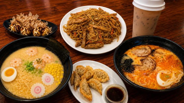 A spread of Japanese dishes on a wooden table at Tasty Oishi, featuring shrimp ramen, spicy pork ramen, chicken hibachi noodles, takoyaki topped with bonito flakes, crispy gyoza with dipping sauce, and an iced coffee.
