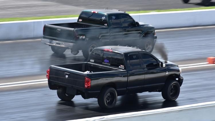 Two diesel pickup trucks race side by side on a drag strip, releasing black smoke as they accelerate.