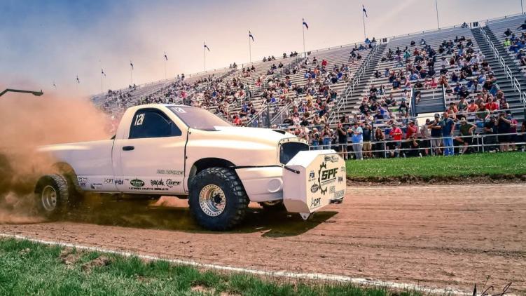 Modified diesel truck competes in a sled pull, kicking up dirt as a large crowd watches from the grandstands.