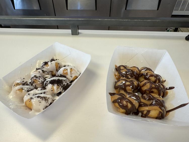 Reese’s PB & Chocolate and Cookies N’ Cream Mini Donuts at Hometown Mini Donuts Reese’s PB & Chocolate and Cookies N’ Cream Mini Donuts at Hometown Mini DonutsTwo trays of specialty mini donuts from Hometown Mini Donuts displayed side by side on a white counter. One tray features Cookies N’ Cream mini donuts topped with vanilla icing and crushed chocolate cookies, while the other features Reese’s-inspired mini donuts drizzled with chocolate and peanut butter sauce.