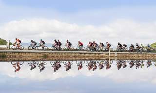 Um grupo de ciclistas pedala por uma estrada junto às salinas de Ibiza durante um evento desportivo, com os reflexos dos participantes na água, montanhas em segundo plano e um céu parcialmente nublado.