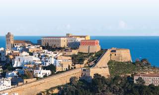Vista panorâmica de Dalt Vila, a cidade amuralhada de Ibiza, Património Mundial da UNESCO, com as suas muralhas renascentistas, a catedral a dominar a paisagem urbana, casas brancas na sua base e o Mar Mediterrâneo como pano de fundo.