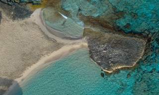 A praia de Ibiza vista do céu, com as águas turquesas e transparentes do Mar Mediterrâneo e uma grande rocha que se estende desde a areia até ao mar.