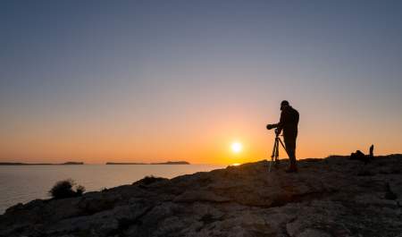 Um homem com uma câmara profissional fotografa o pôr do sol em Ibiza a partir da costa, com vista para o mar.