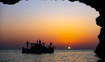 Um grupo de amigos acena de um barco contra a luz, num pôr-do-sol alaranjado em pleno mar em Ibiza.