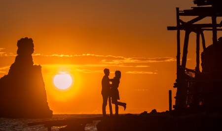 Um casal abraça-se numa praia de Ibiza durante um belo pôr-do-sol, com o céu alaranjado e as suas silhuetas escuras iluminadas por trás.