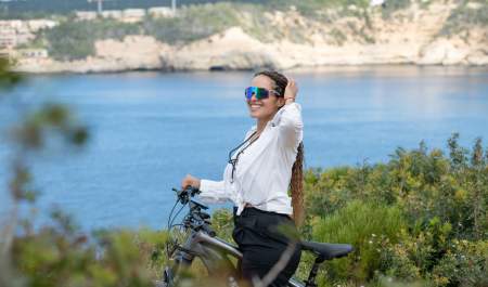 Uma menina com óculos de sol e de pé sozinha está a andar de bicicleta à beira-mar numa área natural verde.