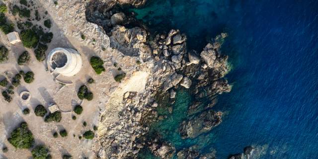 Vista aérea de la Torre des Savinar, una histórica torre de defensa situada sobre un acantilado de Ibiza, rodeada de costa rocosa y aguas cristalinas del mar Mediterráneo en diferentes tonalidades de azul.