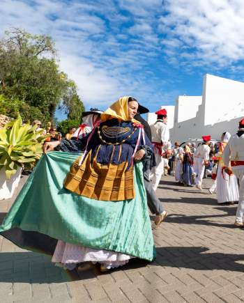 A group of dancers in traditional Ibizan costumes perform the *ball pagès* in front of a white church decorated with pennants during a local festival, surrounded by residents and visitors under a blue sky.