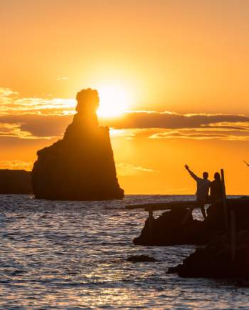 Atardecer en la costa de Ibiza con una pareja contemplando el mar desde un embarcadero. El sol se oculta tras una formación rocosa junto al horizonte, tiñendo el cielo de tonos dorados y anaranjados, mientras las siluetas de las personas y las construcciones costeras se recortan sobre el paisaje mediterráneo.