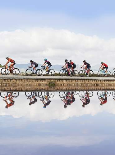 Um grupo de ciclistas pedala por uma estrada junto às salinas de Ibiza durante um evento desportivo, com os reflexos dos participantes na água, montanhas em segundo plano e um céu parcialmente nublado.